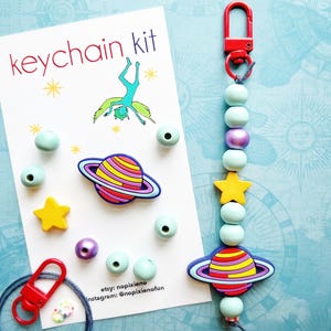 May include: A keychain kit with a red clasp, light blue beads, a yellow star, and a planet-shaped charm. The kit includes a card that says "keychain kit" with a drawing of a person doing a handstand. Additional beads and charms are scattered around the card.