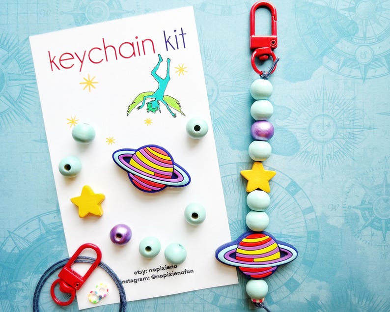 May include: A keychain kit with a red clasp, light blue beads, a yellow star, and a planet-shaped charm. The kit includes a card that says "keychain kit" with a drawing of a person doing a handstand. Additional beads and charms are scattered around the card.