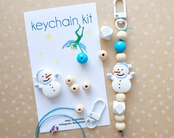 May include: A DIY keychain kit featuring a snowman charm, wooden beads, a blue bead, a white hexagonal bead, and a silver clasp. The kit also includes a card with the text "keychain kit" and social media handles for the seller.