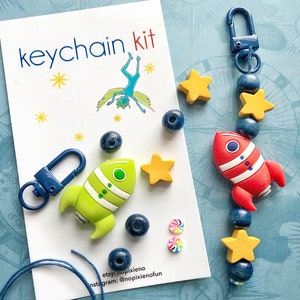 May include: A DIY keychain kit with a blue and white rocket, yellow stars, and blue beads. The kit also includes a blue metal keychain clasp, green and white rocket charm, and a card that says "keychain kit".