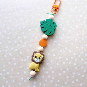 May include: A colorful wooden toy keychain with a green monstera leaf, orange and white beads, and a brown and yellow lion charm.