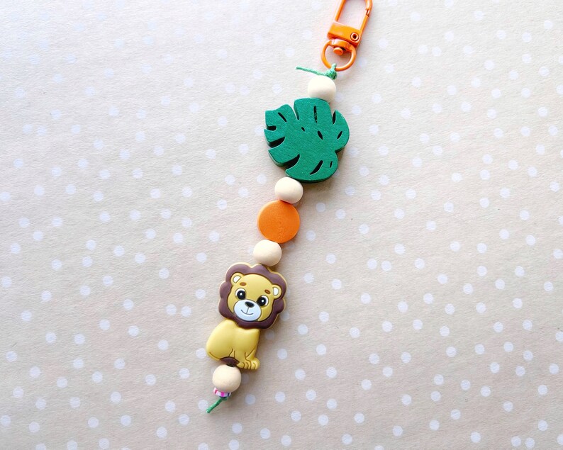 May include: A colorful wooden toy keychain with a green monstera leaf, orange and white beads, and a brown and yellow lion charm.