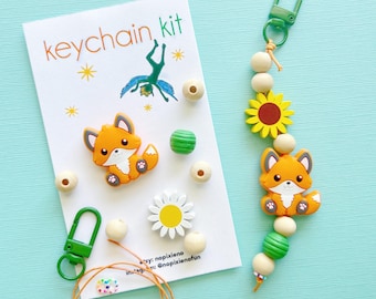 May include: A keychain kit with a cute orange fox charm, wooden beads, a green clasp, and a sunflower charm. The kit includes a card that says "keychain kit" with a drawing of a person doing a flip. The kit is on a light blue background.