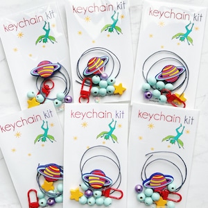 May include: Several keychain kits are displayed, each packaged in a white card with the text "keychain kit". Each kit includes a planet-shaped charm, beads, a star charm, and a red clasp. The kits have a space theme.