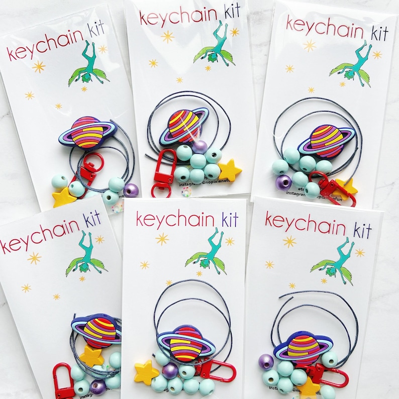 May include: Several keychain kits are displayed, each packaged in a white card with the text "keychain kit". Each kit includes a planet-shaped charm, beads, a star charm, and a red clasp. The kits have a space theme.