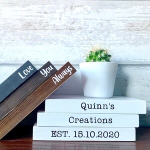 May include: A stack of four white books with black text. The top book says "Quinn's Creations" and the bottom book says "EST. 15.10.2020". Three wooden blocks with the words "Love", "You", and "Always" are stacked on top of the books. A small potted cactus is to the right of the books.