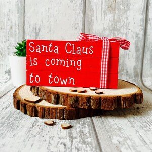 May include: Red wooden blocks stacked on top of each other with the words "Santa Claus is coming to town" written in white. The blocks are sitting on a wooden slice.