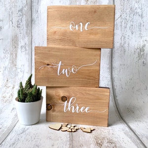 May include: Three wooden signs stacked on top of each other. The signs are painted brown and have white calligraphy writing. The top sign says "one", the middle sign says "two", and the bottom sign says "three".