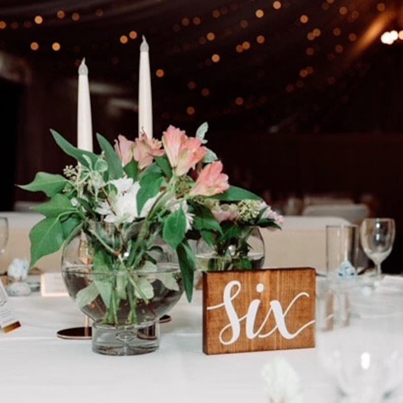 May include: A wooden table number with the word "six" written in white calligraphy. The table number is on a white tablecloth with a floral centerpiece in a clear glass vase.