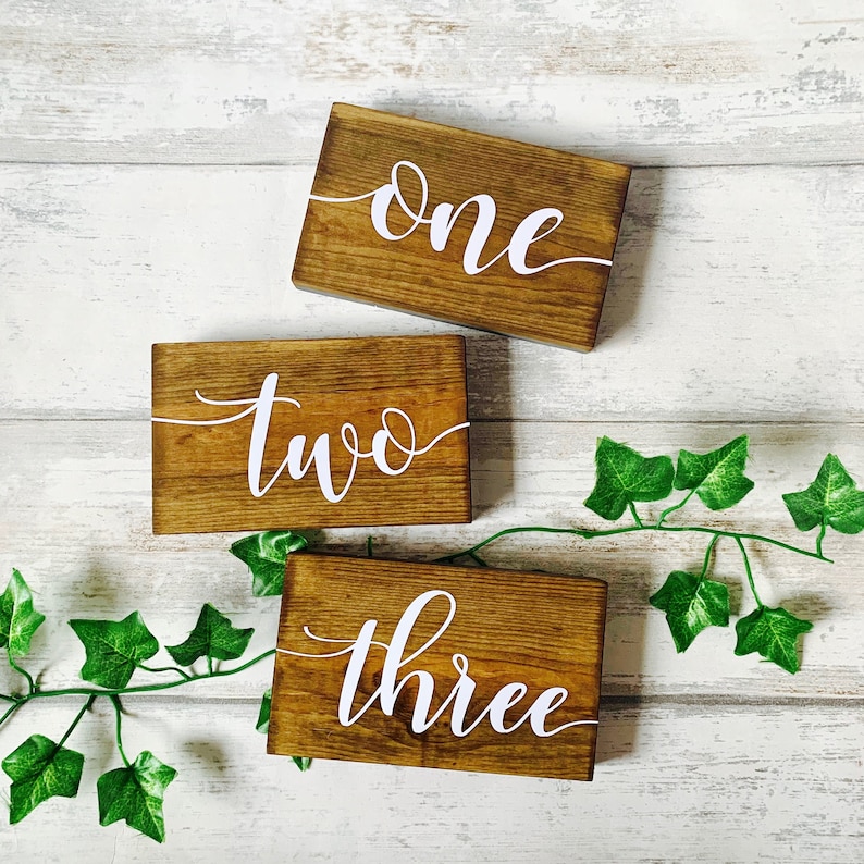 May include: Three wooden signs with the words "one", "two", and "three" written in white cursive font. The signs are on a white wooden background with green ivy.