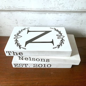 May include: Three white wooden blocks stacked on top of each other. The top block has a black letter "Z" with a wreath design around it. The middle block says "The Nelsons" and the bottom block says "EST. 2010".