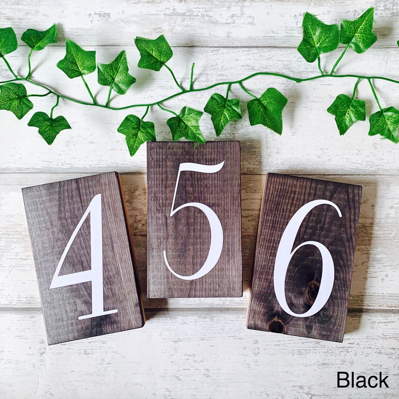 May include: Three wooden blocks with the numbers 4, 5, and 6 painted in white on a dark brown background. The blocks are arranged in a row on a white wooden surface. The word "Black" is written in white letters on the bottom right corner of the image.