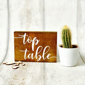 May include: A rustic wooden sign with the words "top table" written in white paint. The sign is sitting on a white wooden surface.