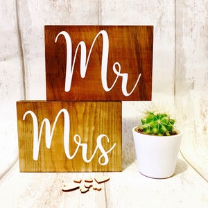 May include: Two wooden signs with the words "Mr." and "Mrs." in white lettering. The signs are on a white wooden background with a small potted cactus in the foreground.