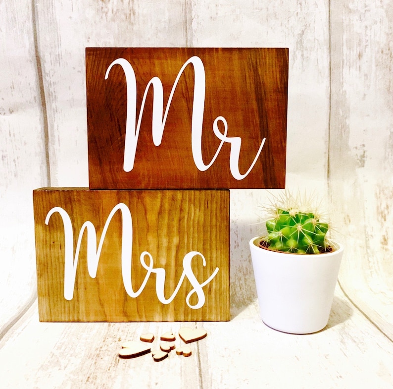 May include: Two wooden signs with the words "Mr." and "Mrs." in white lettering. The signs are on a white wooden background with a small potted cactus in the foreground.