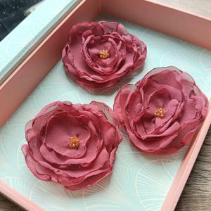 Pode incluir: Três flores de tecido em rosa seco com pétalas sobrepostas semitransparentes e um centro de pequenas contas douradas. Cada flor apresenta bordas delicadas ligeiramente desfiadas e uma forma arredondada, dispostas numa bandeja rosa.