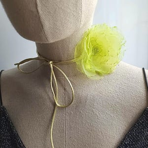 Op de afbeelding: Een choker-halsketting met een limoengroene stoffen bloem, bestaande uit gelaagde, doorschijnende en geplooide bloemblaadjes met een kraal in het midden. De bloem zit vast aan een dun, bijpassend koord dat aan de voorzijde gestrikt wordt.