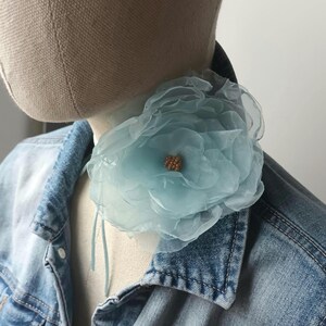 May include: A light blue, layered fabric flower with a cluster of small, gold-colored beads in the center. The flower is attached to a light blue ribbon and is displayed on a denim jacket.