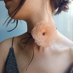 Könnte beinhalten: Eine zarte, pfirsichfarbene Blumen-Choker-Kette mit einem braunen Band. Die Blume besteht aus mehreren Lagen Stoff und hat eine kleine braune Mitte.