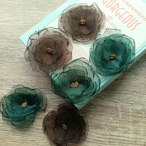 Può includere: Sei abbellimenti floreali in tessuto nei toni del marrone e del verde acqua con centri dorati. I fiori sono stratificati con petali trasparenti. I fiori sono disposti su una scatola azzurra con la parola "GORGEOUS" visibile.