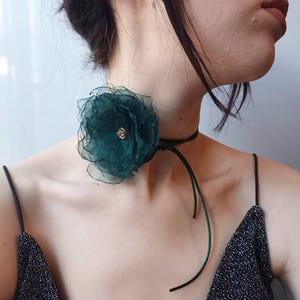 May include: A dark green fabric flower choker necklace with a gold accent. The necklace is tied around the neck of a person wearing a black top.