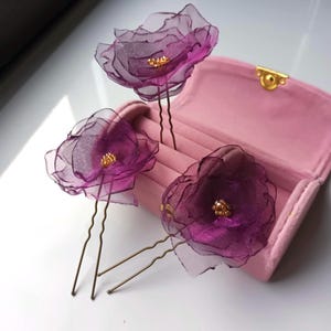 Puede incluir: Tres pinzas para el pelo con forma de flor morada con detalles dorados en una caja de terciopelo rosa.