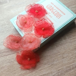 Può includere: Abbellimenti floreali in tessuto trasparente rosso e rosa con centri dorati sono disposti vicino a una scatola azzurra. La scatola ha la scritta "Get Guaranteed Gorgeous". I fiori sono su una superficie di legno.