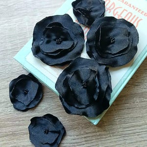 Op de afbeelding: Vijf zwarte stoffen bloemversieringen met kralenmiddens zijn gerangschikt op een lichtblauw boek met de woorden "GET GUARANTEED GORGEOUS" gedrukt op de kaft. De bloemen zijn gemaakt van gelaagde bloemblaadjes.
