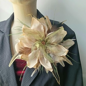 Puede incluir: Un broche floral grande con pétalos de tela color crema en capas, detalles de organza verde oliva transparente y plumas marrones. El centro de la flor está adornado con un grupo de pequeñas cuentas redondas de color bronce metálico.