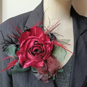Puede incluir: Un broche de rosa de tela en color burdeos intenso, acentuado con hojas verde oscuro y plumas finas negras y rojas. El accesorio floral está prendido en un blazer gris carbón a rayas finas sobre una prenda superior negra en un maniquí.