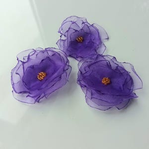 Op de afbeelding: Drie handgemaakte paarse stoffen bloemen met gelaagde bloemblaadjes en een gouden kralenmiddelpunt. De bloemen zijn gerangschikt op een wit oppervlak, waarbij hun delicate, transparante textuur en levendige kleur worden getoond.
