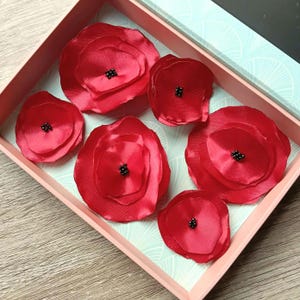 May include: Six red fabric flower decorations with black centres, presented in a pink box with a light blue patterned interior. The flowers, of varying sizes and shapes, suggest a handmade craft item.