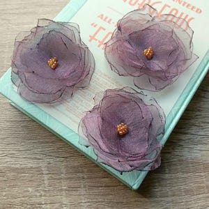 Puede incluir: Tres adornos florales de tela morada hechos a mano con centros dorados descansan sobre una caja azul claro y blanca. Las flores tienen múltiples capas de tela transparente, creando una apariencia delicada y texturizada. La caja tiene el texto "GET GUARANTEED GORGEOUS".