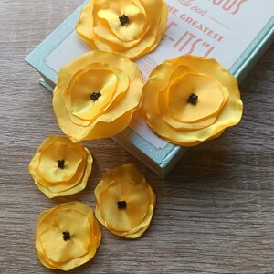 May include: Six yellow fabric flower embellishments with black centers are arranged on a wooden surface next to a book. The book has the text "GET GUARANTEED GORGEOUS with our GREATEST -ITS!" printed on the cover.