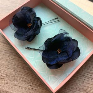 Pode incluir: Duas presilhas de cabelo de flor azul marinho com centros de contas douradas repousam numa caixa decorativa. A caixa tem uma borda rosa e um interior com padrão azul claro e branco. As flores são feitas de tecido em camadas e material transparente.
