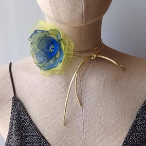 Può includere: Una collana girocollo con un grande fiore in tessuto a strati nei toni del giallo e del blu. Il fiore presenta un centro blu scuro con piccoli accenti verdi. Il girocollo è fissato con un sottile cordino color oro metallizzato annodato davanti.