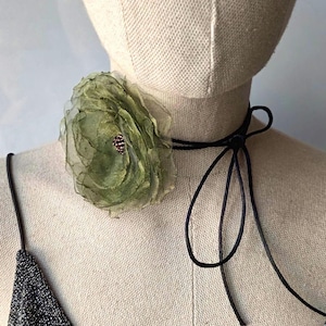 Könnte beinhalten: Eine Halskette mit einer großen, mehrlagigen Stoffblume in Olivgrün, die in der Mitte ein kleines, dunkles und strukturiertes Detail aufweist. Die Blume ist an einem dünnen, schwarzen Band befestigt, das seitlich gebunden ist und auf einem beigen Schneiderbüsten-Torso liegt.
