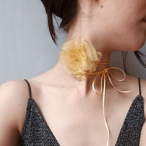 Può includere: Un delicato girocollo dorato con un grande fiore giallo in tessuto. Il fiore è a strati e ha una texture leggermente arricciata.