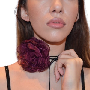 Puede incluir: Un collar choker con una flor de tela de color morado oscuro y lazos de cinta negra. La flor es grande y tiene una textura arrugada.