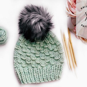 May include: A light green knitted beanie with a black and white faux fur pom pom. The beanie is made with chunky yarn and has a textured stitch pattern. The beanie is lying on a white surface with a ball of yarn and knitting needles.