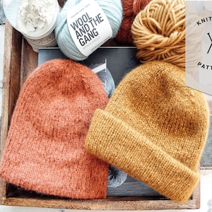 May include: Two knitted hats, one in a rust orange color and one in a mustard yellow color, are displayed in a wooden tray. The hats are made with a chunky yarn and have a simple, slouchy design. The tray also contains balls of yarn in various colors, including blue, yellow, and orange.