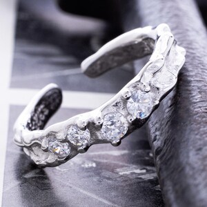 Könnte beinhalten: Ein silberfarbener Ring mit einer strukturierten, organischen Gestaltung. Der Ring ist mit mehreren klaren, runden Edelsteinen besetzt. Der Ring ist unten offen, was eine Größenanpassung ermöglicht. Präsentiert auf einem schwarz-weiß gemusterten Hintergrund.