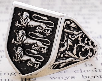 May include: A silver signet ring featuring a shield-shaped face with three embossed lions stacked vertically against a dark background. The sides of the ring are decorated with intricate, raised silver scrollwork and floral patterns.