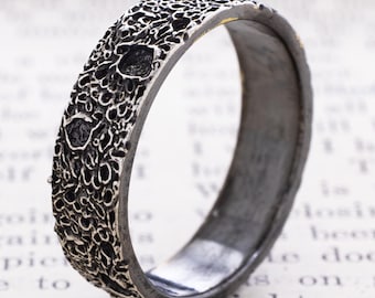May include: A silver-toned metal ring featuring a highly textured, cratered surface that mimics the appearance of a lunar landscape. The band has a dark, oxidized finish within the recessed areas and a smooth, polished interior surface.