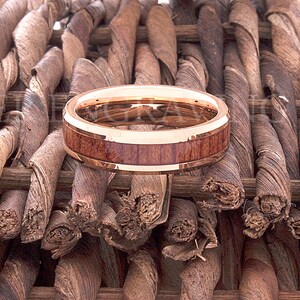 May include: A rose gold tungsten carbide ring with a brown wood inlay. The ring is sitting on a woven brown background.