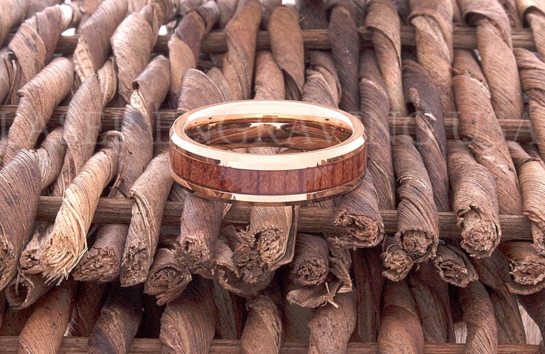 May include: A rose gold tungsten carbide ring with a brown wood inlay. The ring is sitting on a woven brown background.
