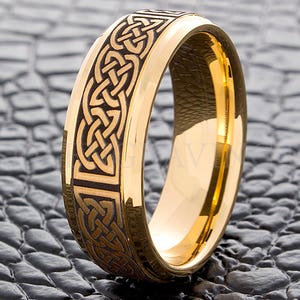 May include: A gold-colored ring with a Celtic knot design etched into a dark band. The ring has a polished, reflective surface and is set against a textured, dark gray background.