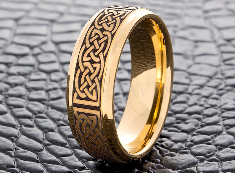 May include: A gold-colored ring with a Celtic knot design etched into a dark band. The ring has a polished, reflective surface and is set against a textured, dark gray background.