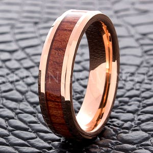 May include: A rose gold tungsten carbide ring with a brown wood inlay. The ring has a beveled edge and a polished finish.