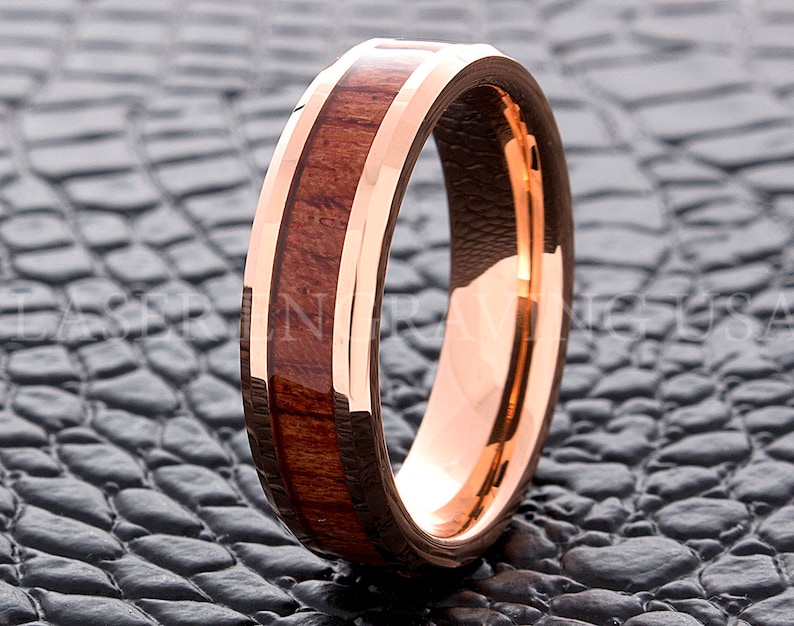 May include: A rose gold tungsten carbide ring with a brown wood inlay. The ring has a beveled edge and a polished finish.